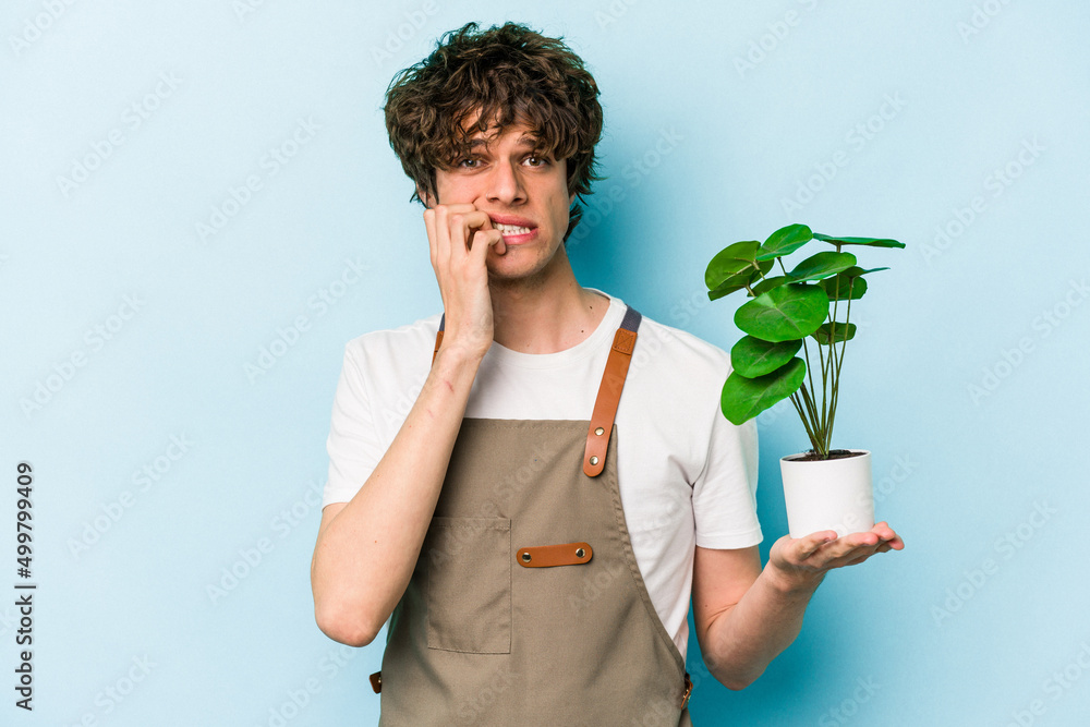 Young gardener caucasian man holding a plant isolated on blue background biting fingernails, nervous and very anxious. Young gardener caucasian man holding a plant isolated on blue background biting fingernails, nervous and very anxious.