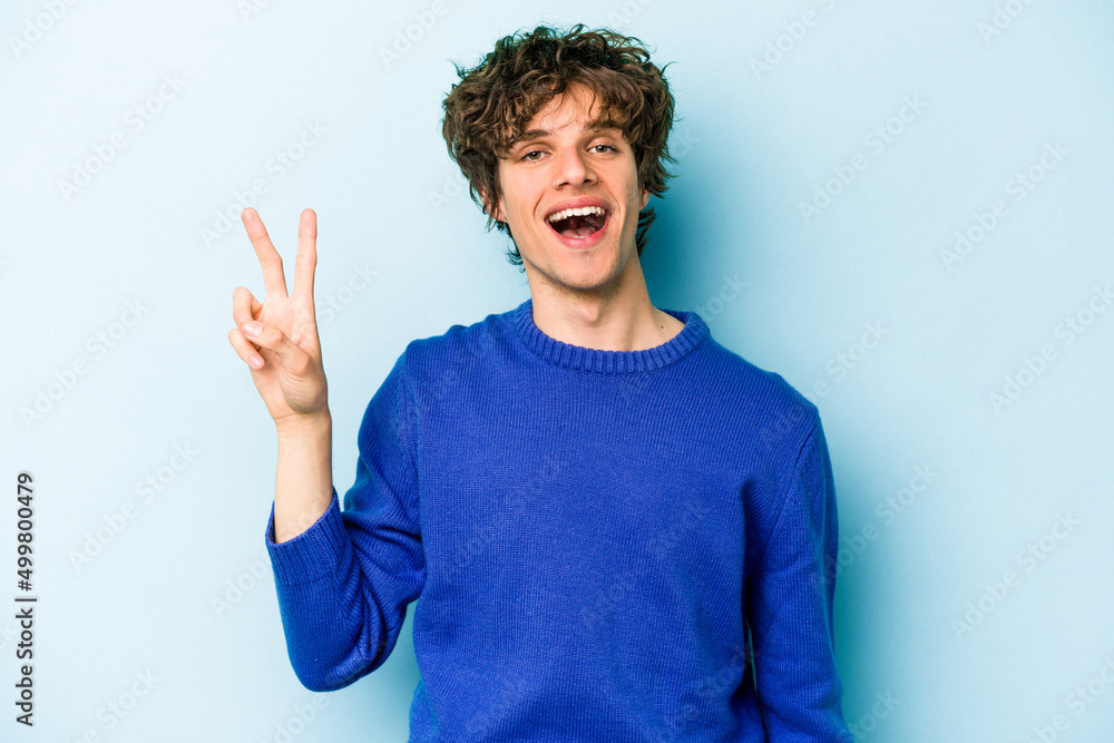 Young caucasian man isolated on blue background joyful and carefree showing a peace symbol with fingers. Young caucasian man isolated on blue background joyful and carefree showing a peace symbol with fingers.