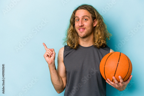 Young caucasian man playing basketball isolated on blue background smiling and pointing aside, showing something at blank space.