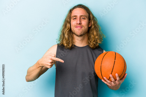 Young caucasian man playing basketball isolated on blue background person pointing by hand to a shirt copy space, proud and confident