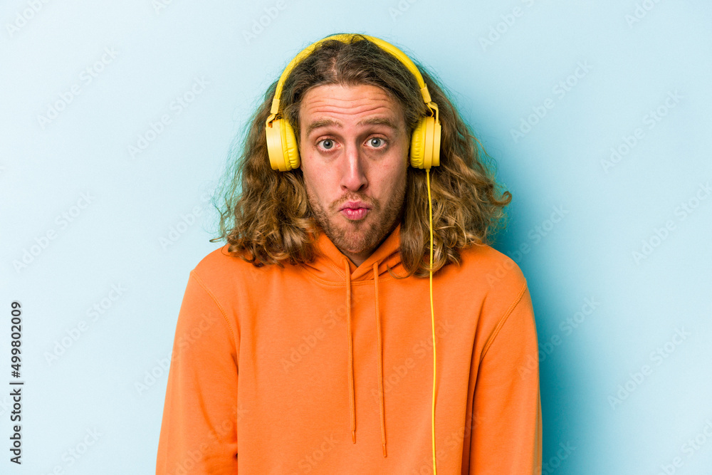 Fototapeta premium Young caucasian man listening to music isolated on blue background shrugs shoulders and open eyes confused.