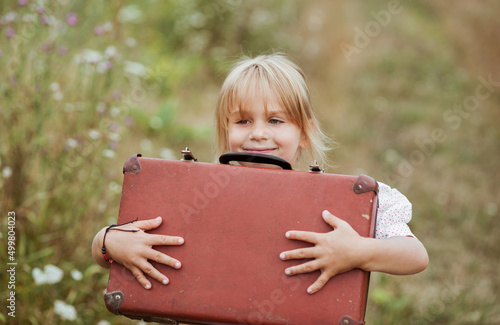 Portrait of a cheerful little girl holding her old suitcases and smiling in the countryside at summer time with a copy space for text