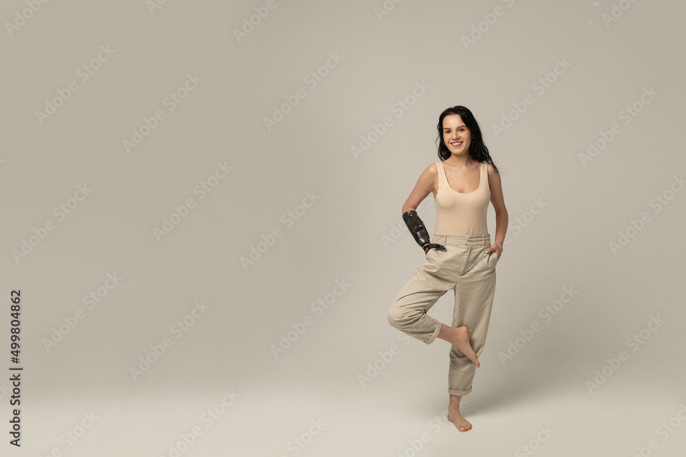 Beauty disabled Woman with prosthetic arm, artificial hand looking at camera, smiling, stands on one leg in crane pose isolated on beige background. Beauty variety. Positive. Copy space