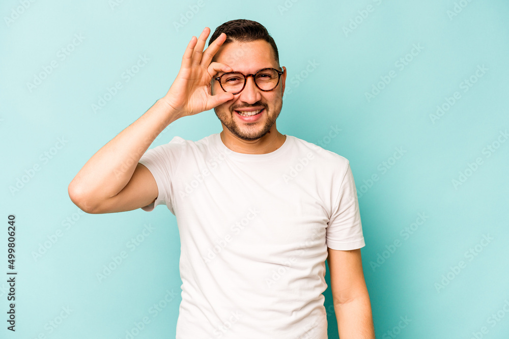 Young hispanic man isolated on blue background excited keeping ok gesture on eye. Young hispanic man isolated on blue background excited keeping ok gesture on eye.