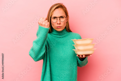 Young caucasian woman holding tupperware isolated on pink background showing fist to camera, aggressive facial expression. photo