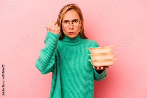 Cuadro en lienzo Young caucasian woman holding tupperware isolated on pink background showing fist to camera, aggressive facial expression