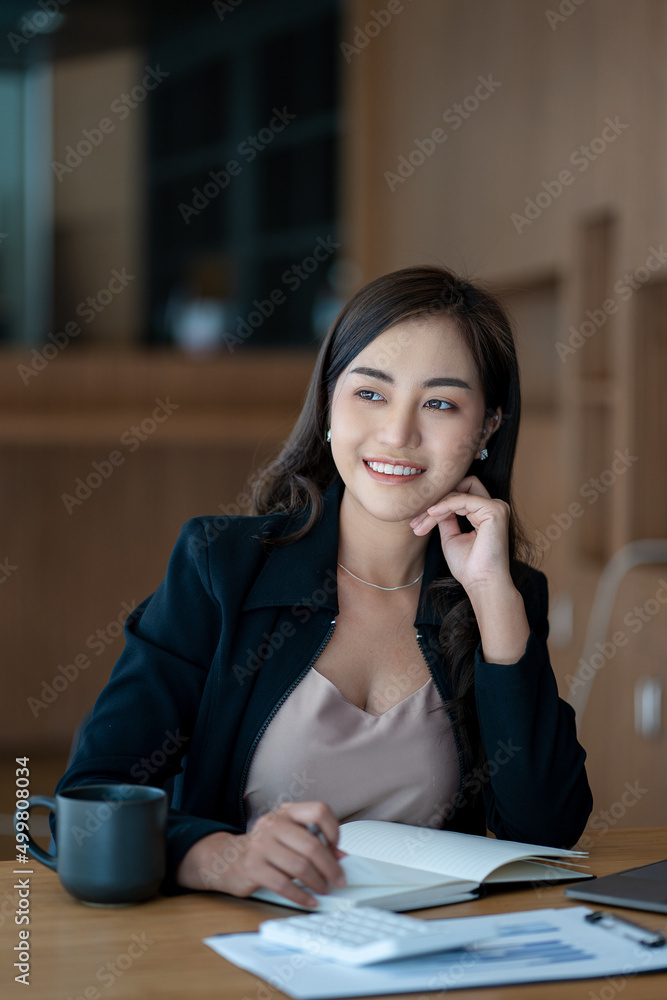 Smiling Asian businesswoman working on paper and holding pen and laptop sitting on a chair at modern home studio. Mobile workers at work and calculators Finance or Accounting Concepts Smiling Asian businesswoman working on paper and holding pen and laptop sitting on a chair at modern home studio. Mobile workers at work and calculators Finance or Accounting Concepts
