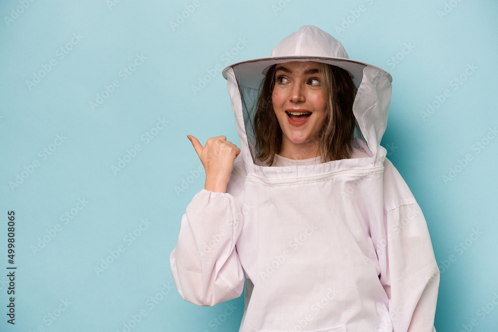 Young caucasian beekeeper woman isolated on blue background points with thumb finger away, laughing and carefree. Young caucasian beekeeper woman isolated on blue background points with thumb finger away, laughing and carefree.
