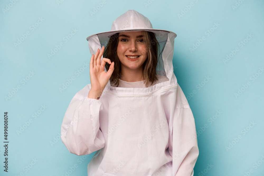 Young caucasian beekeeper woman isolated on blue background cheerful and confident showing ok gesture. Young caucasian beekeeper woman isolated on blue background cheerful and confident showing ok gesture.