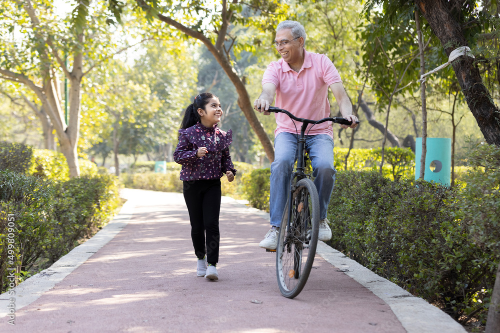 Cheerful senior man riding bicycle while granddaughter running at park