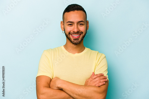 Young hispanic man isolated on blue background who feels confident, crossing arms with determination.