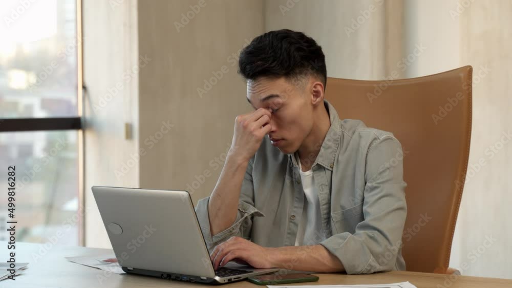 Tired Asian man rubs sleepy eyes working on laptop in office. Young employee types text on keyboard sitting at wooden table near bright window closeup Tired Asian man rubs sleepy eyes working on laptop in office. Young employee types text on keyboard sitting at wooden table near bright window closeup