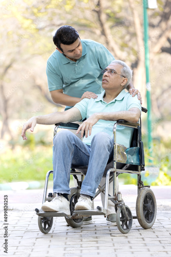 Young man assisting a senior man in a wheelchair at park Young man assisting a senior man in a wheelchair at park