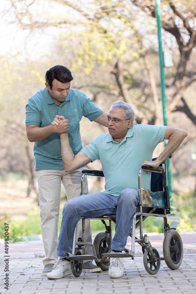Young man assisting a senior man in a wheelchair at park Young man assisting a senior man in a wheelchair at park
