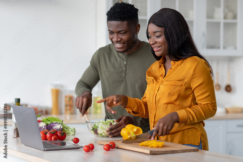 Cheerful Black Spouses Cooking Lunch In Kitchen And Checking Recipes On Laptop