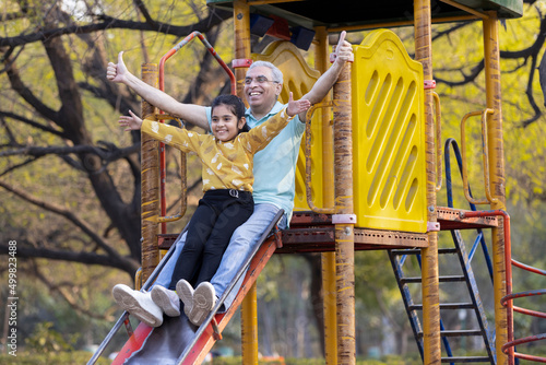 Senior man with granddaughter having fun while sliding in playground at park