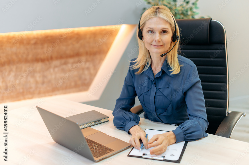Obraz premium Portrait elegant caucasian attractive mature blonde woman with headset, sitting in office, working as operator of call center, support service, financial mentor, looking at camera, smiling friendly