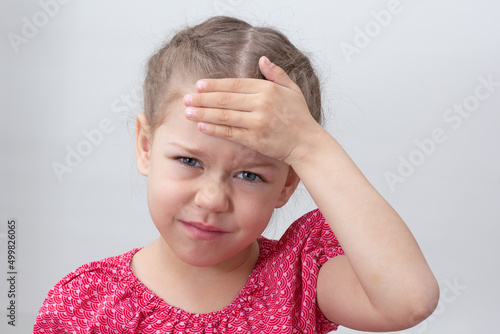 Child with headache holding hand on forehead on white background caucasian little girl 5-6 years in red looking at camera photo