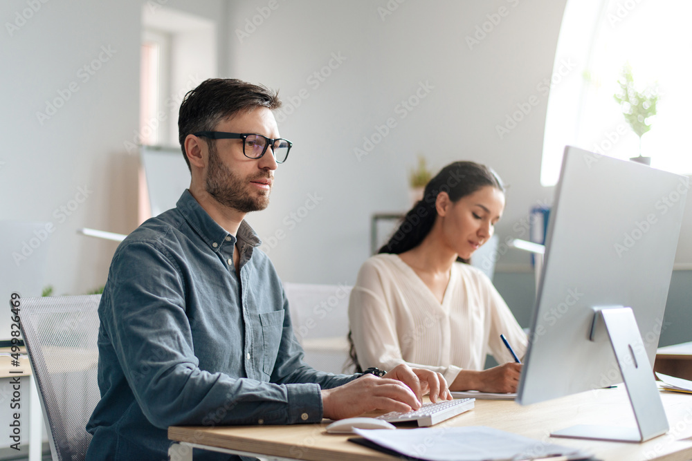 Focused Caucasian man using computer, working in modern office with his colleague, copy space Focused Caucasian man using computer, working in modern office with his colleague, copy space