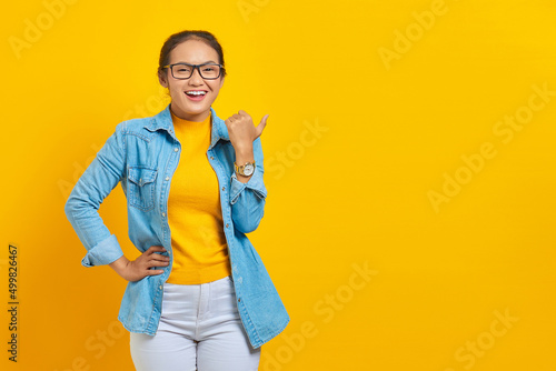 Portrait of cheerful young Asian woman student in denim clothes pointing finger in copy space, showing advertising products isolated on yellow background. Education in college university concept © Bangun Stock Photo