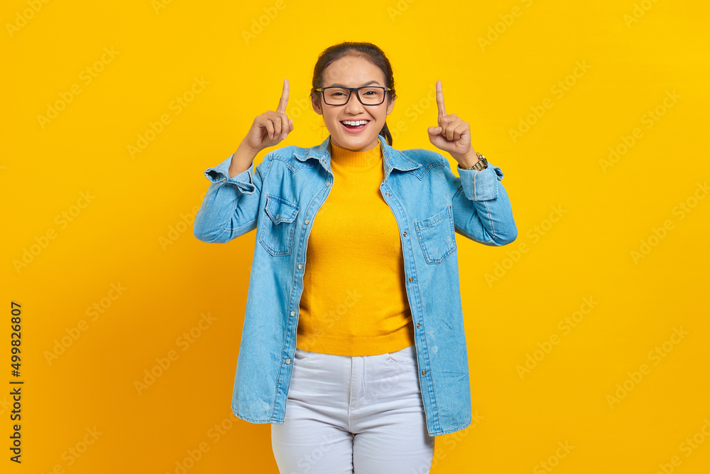 Portrait of smiling young Asian woman student in denim clothes pointing fingers up, inviting customers to special event isolated on yellow background. Education in college university concept Portrait of smiling young Asian woman student in denim clothes pointing fingers up, inviting customers to special event isolated on yellow background. Education in college university concept