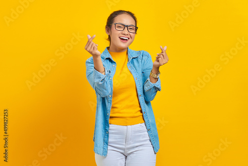 Portrait of smiling young Asian woman student in denim clothes showing korean heart with two fingers crossed isolated on yellow background. People sincere emotions lifestyle concept photo
