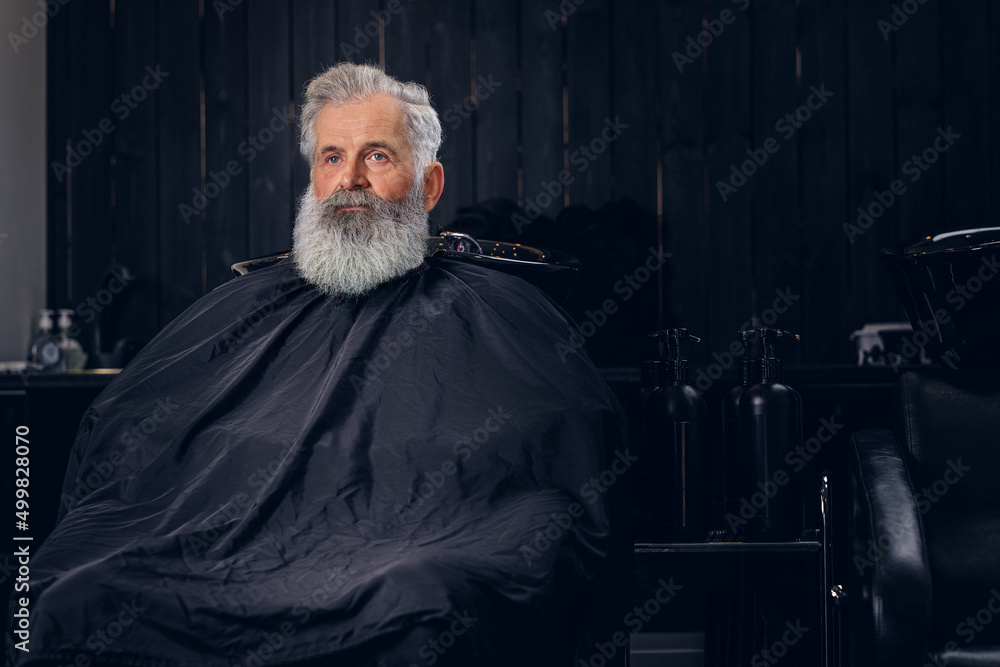 Shot of fashionable old hipster with long beard and peignoir in dark barber shop.
