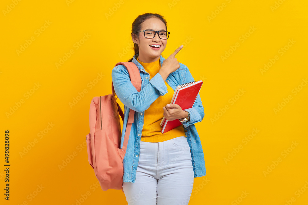 Portrait of smiling young Asian woman student in casual clothes with backpack holding book and pointing aside with finger isolated on yellow background. Education in college university concept Portrait of smiling young Asian woman student in casual clothes with backpack holding book and pointing aside with finger isolated on yellow background. Education in college university concept