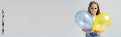 child looking at camera while holding blue and yellow balloons isolated on grey, banner. © LIGHTFIELD STUDIOS