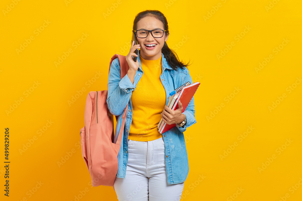 Portrait of cheerful young Asian woman student in denim clothes with backpack talking on mobile phone and holding book isolated on yellow background Portrait of cheerful young Asian woman student in denim clothes with backpack talking on mobile phone and holding book isolated on yellow background