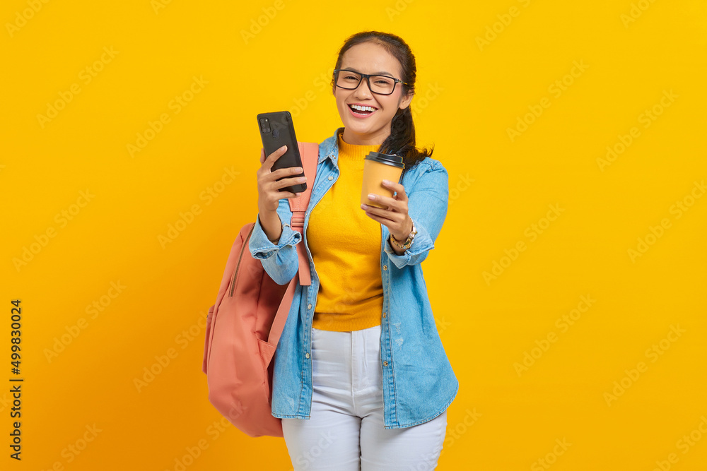 Portrait of cheerful young Asian woman student in denim clothes with backpack holding cup of coffee and smartphone isolated on yellow background Portrait of cheerful young Asian woman student in denim clothes with backpack holding cup of coffee and smartphone isolated on yellow background