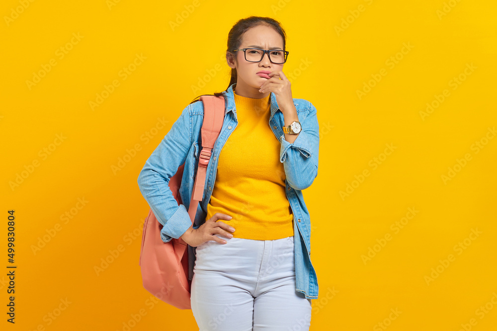 Beautiful young Asian student in denim outfit with backpack and looking serious thinking about question isolated on yellow background. Education in high school university college concept Beautiful young Asian student in denim outfit with backpack and looking serious thinking about question isolated on yellow background. Education in high school university college concept