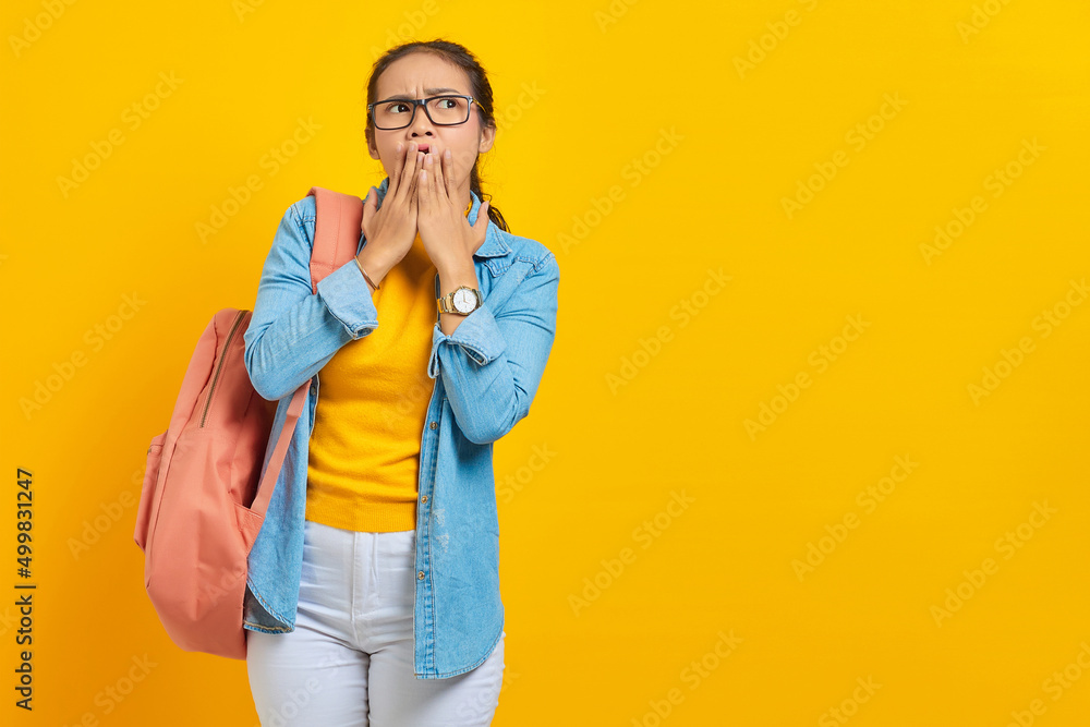 Surprised young Asian student in denim clothes with backpack covering mouth with hands isolated on yellow background. Education in high school university college concept Surprised young Asian student in denim clothes with backpack covering mouth with hands isolated on yellow background. Education in high school university college concept