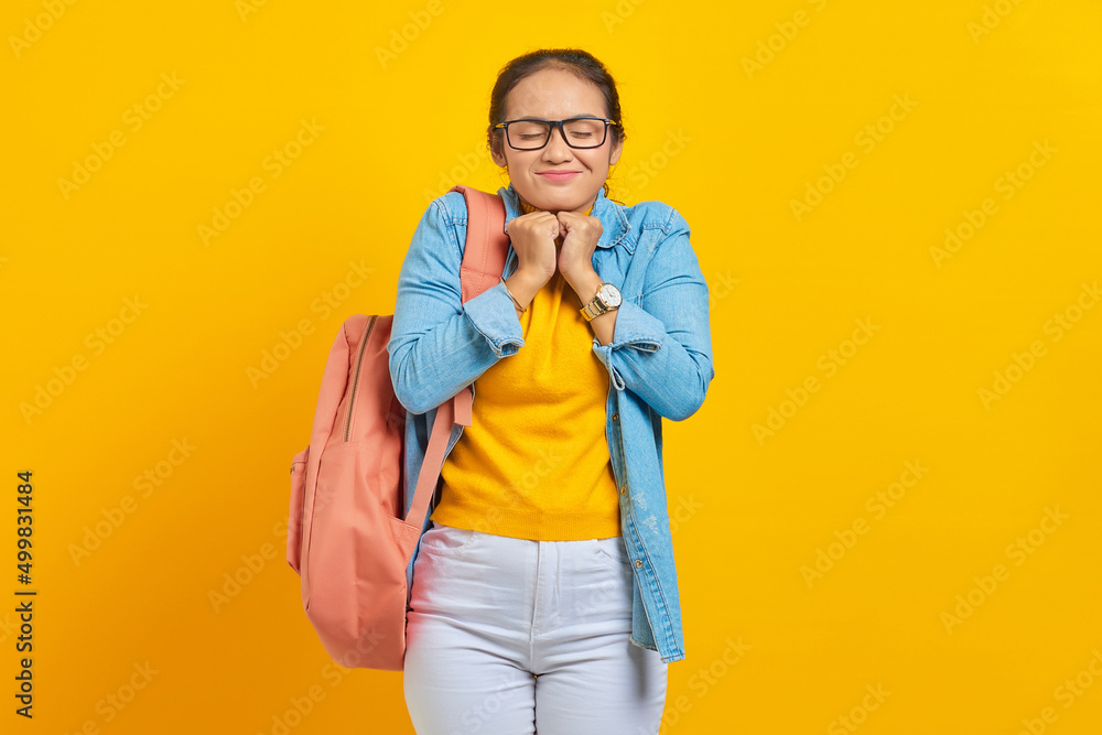 Excited young Asian student in denim clothes with backpack keeps hands under chin, remembers pleasant moment isolated on yellow background. Education in high school university college concept Excited young Asian student in denim clothes with backpack keeps hands under chin, remembers pleasant moment isolated on yellow background. Education in high school university college concept