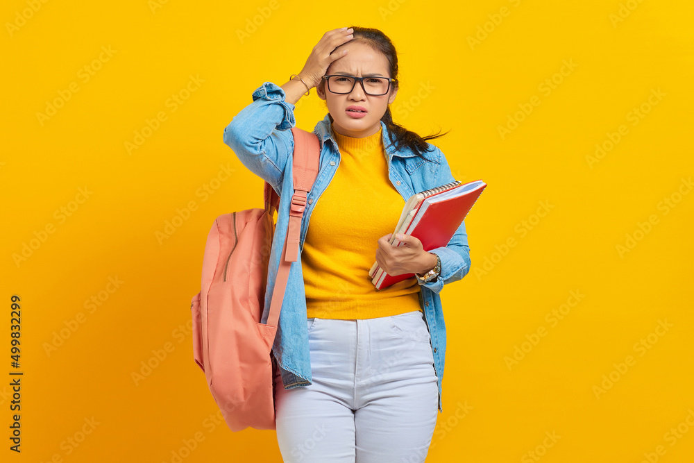 Beautiful young Asian college student in denim outfit with backpack holding notebook and looking dizzy with headache isolated on yellow background. Education in high school university college concept Beautiful young Asian college student in denim outfit with backpack holding notebook and looking dizzy with headache isolated on yellow background. Education in high school university college concept