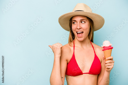 Young caucasian woman wearing a bikini and holding an ice cream isolated on blue background points with thumb finger away, laughing and carefree.