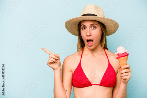 Young caucasian woman wearing a bikini and holding an ice cream isolated on blue background pointing to the side