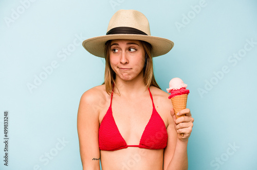 Young caucasian woman wearing a bikini and holding an ice cream isolated on blue background confused, feels doubtful and unsure.
