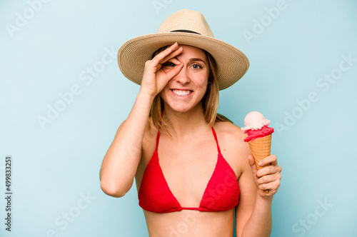 Young caucasian woman wearing a bikini and holding an ice cream isolated on blue background excited keeping ok gesture on eye.