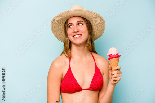 Young caucasian woman wearing a bikini and holding an ice cream isolated on blue background looks aside smiling, cheerful and pleasant.