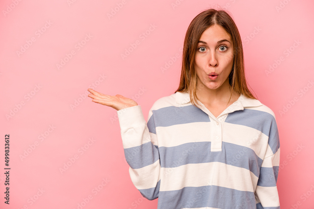 Young caucasian woman isolated on pink background impressed holding copy space on palm. Young caucasian woman isolated on pink background impressed holding copy space on palm.