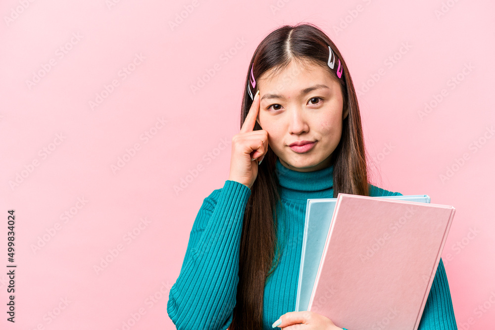 Young asian student woman holding books isolated on pink background pointing temple with finger, thinking, focused on a task. Young asian student woman holding books isolated on pink background pointing temple with finger, thinking, focused on a task.