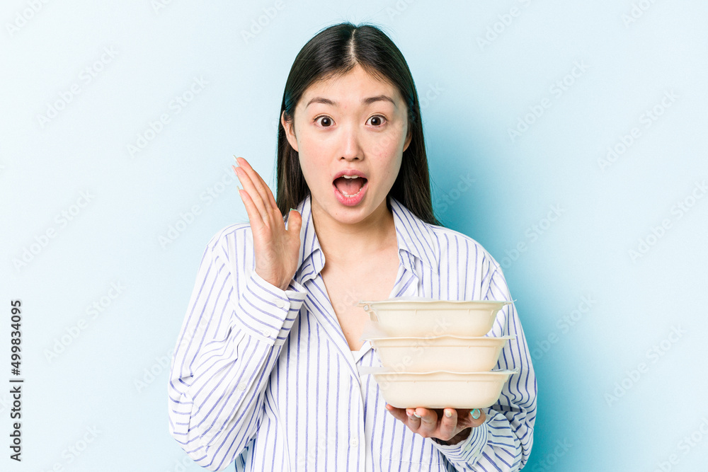 Young asian woman holding Tupperware isolated on blue background surprised and shocked. Young asian woman holding Tupperware isolated on blue background surprised and shocked.