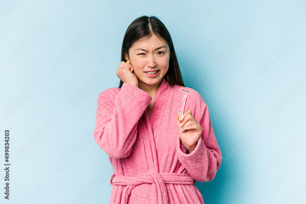 Young asian woman holding razor blade isolated on blue background covering ears with hands. Young asian woman holding razor blade isolated on blue background covering ears with hands.