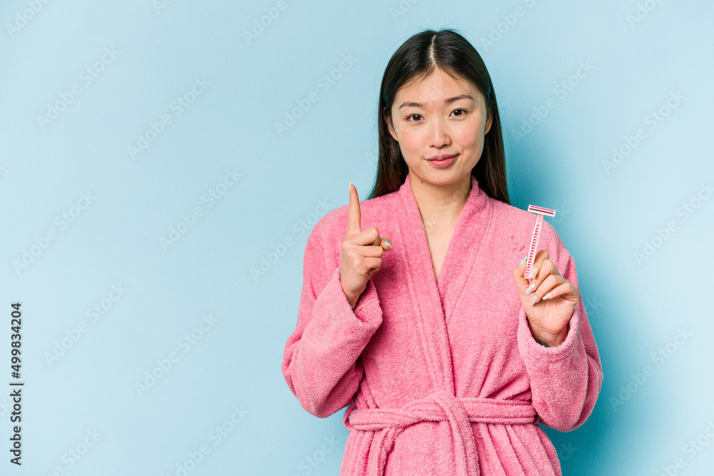 Young asian woman holding razor blade isolated on blue background showing number one with finger. Young asian woman holding razor blade isolated on blue background showing number one with finger.