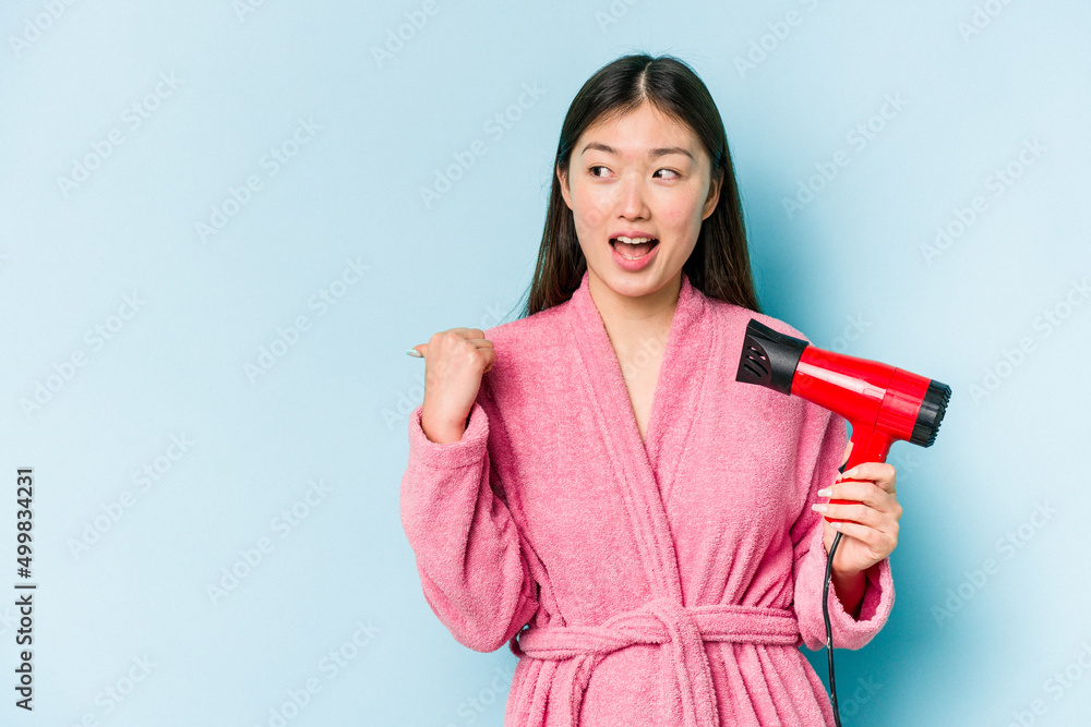 Young asian woman wearing a bathrobe and holding hairdryer isolated on pink background points with thumb finger away, laughing and carefree. Young asian woman wearing a bathrobe and holding hairdryer isolated on pink background points with thumb finger away, laughing and carefree.