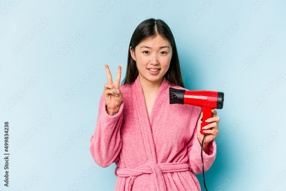 Young asian woman wearing a bathrobe and holding hairdryer isolated on pink background showing number two with fingers. Young asian woman wearing a bathrobe and holding hairdryer isolated on pink background showing number two with fingers.