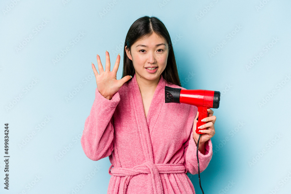 Young asian woman wearing a bathrobe and holding hairdryer isolated on pink background smiling cheerful showing number five with fingers. Young asian woman wearing a bathrobe and holding hairdryer isolated on pink background smiling cheerful showing number five with fingers.