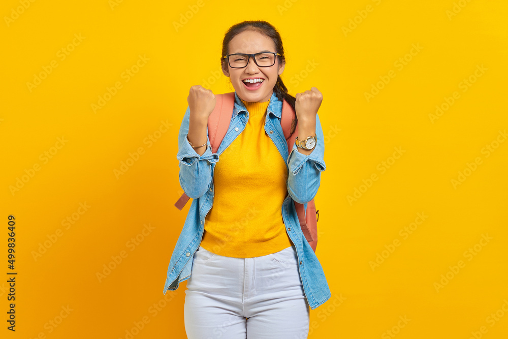 Excited young Asian woman student in denim outfit with backpack standing doing winning gesture celebrating isolated on yellow background. Education in university concept