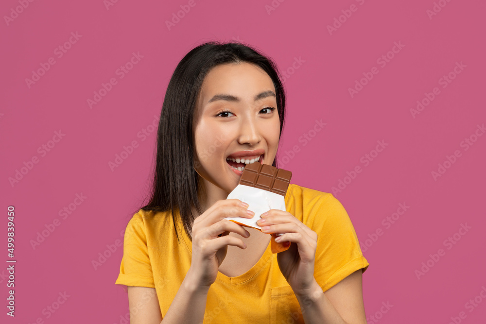 Young korean lady eating tasty chocolate bar, enjoying yummy dessert over violet studio background Young korean lady eating tasty chocolate bar, enjoying yummy dessert over violet studio background